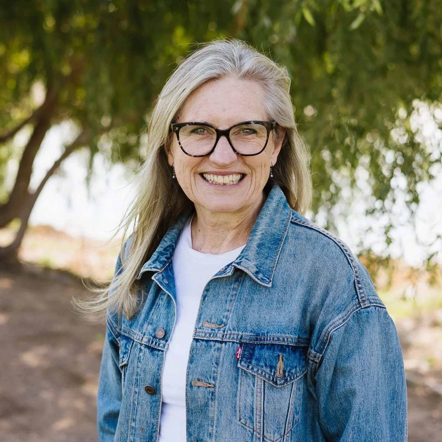 A woman with long gray hair wearing glasses and a denim jacket smiling outdoors with green trees in the background.