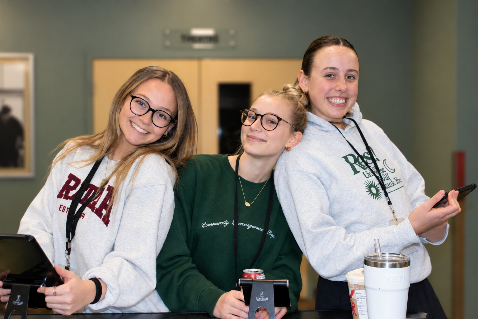A group of young people gathered around a table at a restaurant or café, some smiling and eating, others engaged in conversation, with drinks and food on the table and a large whiteboard in the background.