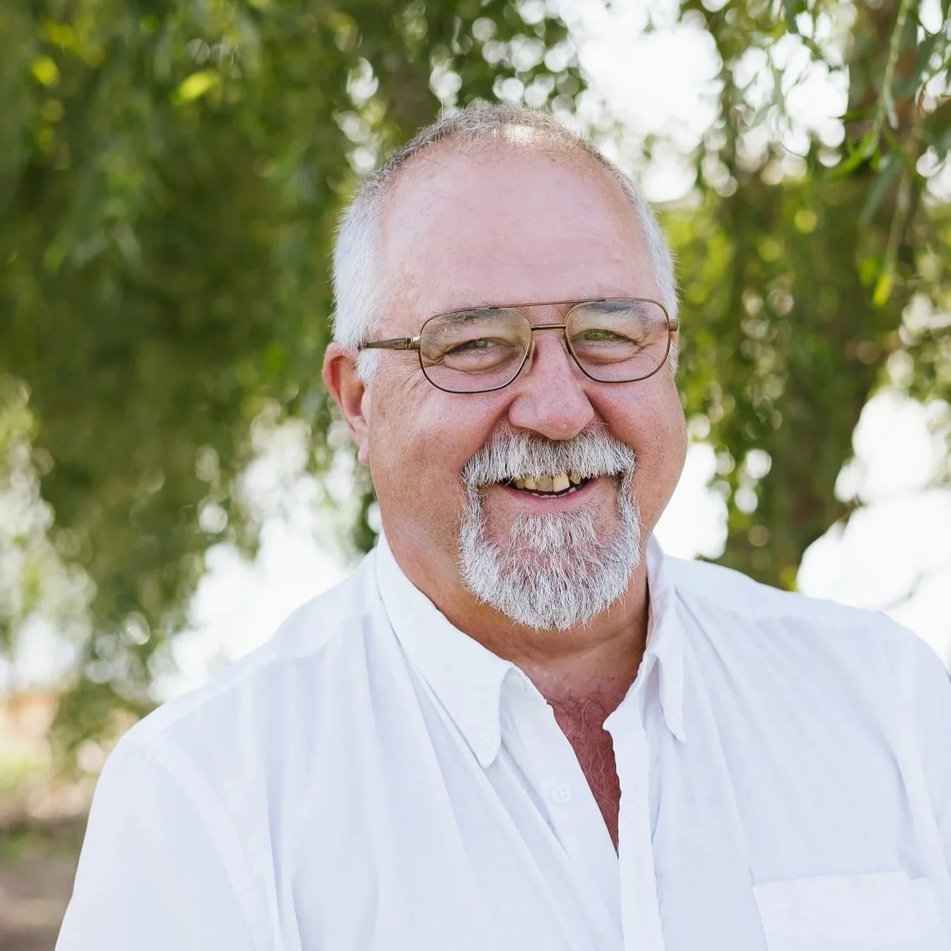 A man with gray hair and a beard wearing a white shirt, smiling outdoors with greenery in the background.