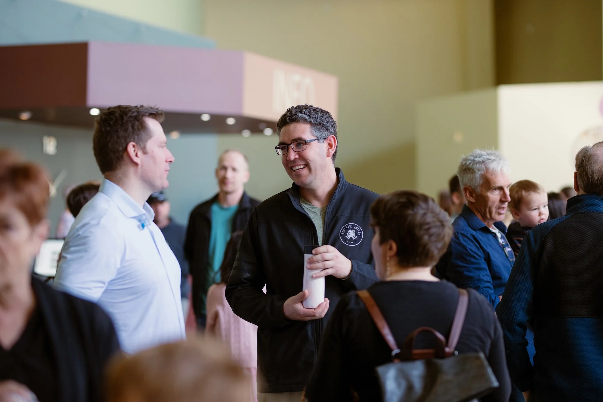 A group of people gathering and talking at an indoor event, with two men in the center engaged in conversation and smiling, surrounded by others of different ages.