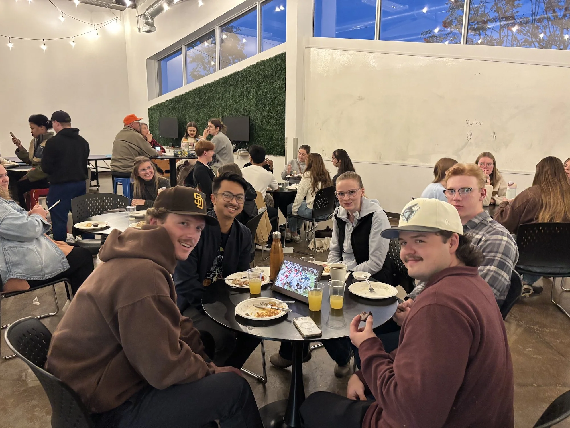 People enjoying a meal together at a social gathering, with some using their phones and others eating and chatting at a table in a well-lit room with large windows.