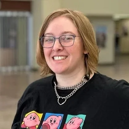 A smiling woman with shoulder-length blonde hair, glasses, wearing a black t-shirt with colorful cartoon characters and a silver chain necklace, standing indoors in a casual setting.