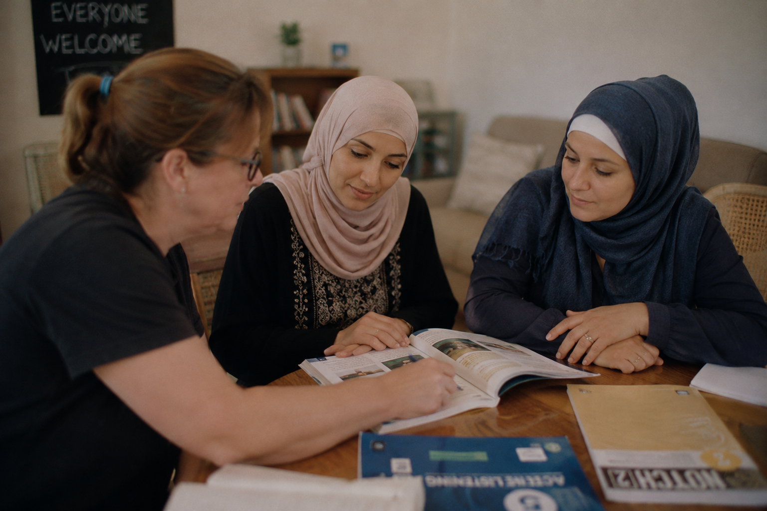 Three women sit at a table looking at a magazine or brochure, with study materials and papers in front of them, in a cozy indoor setting.