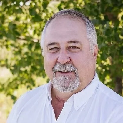 A middle-aged man with short gray hair, a beard, and mustache, wearing a white shirt, outdoors with green foliage in the background.