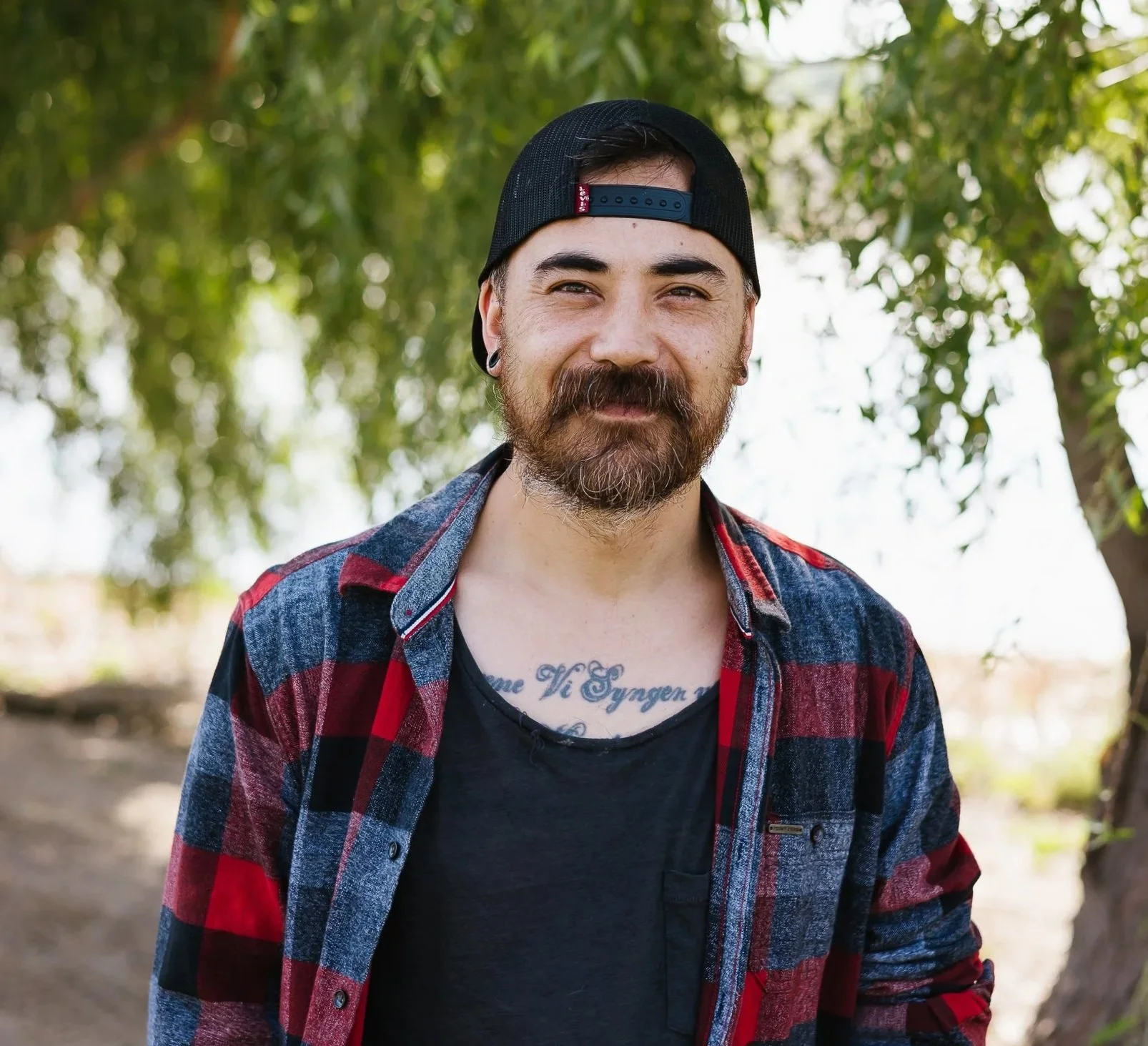 A man outdoors wearing a tan beanie, brown hoodie, and denim jacket, standing in front of trees with fall foliage.