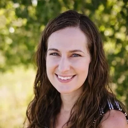 A woman with long brown hair smiling outdoors with green trees in the background.