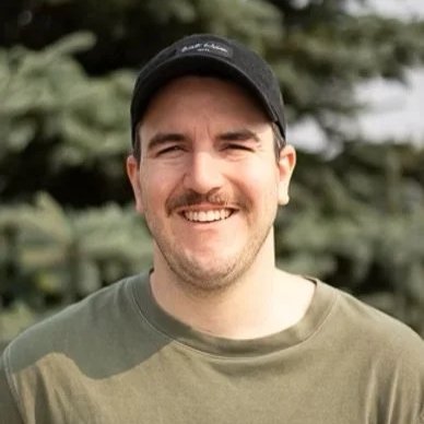A man smiling outdoors, wearing a black cap and a green shirt, with a background of blurred trees.