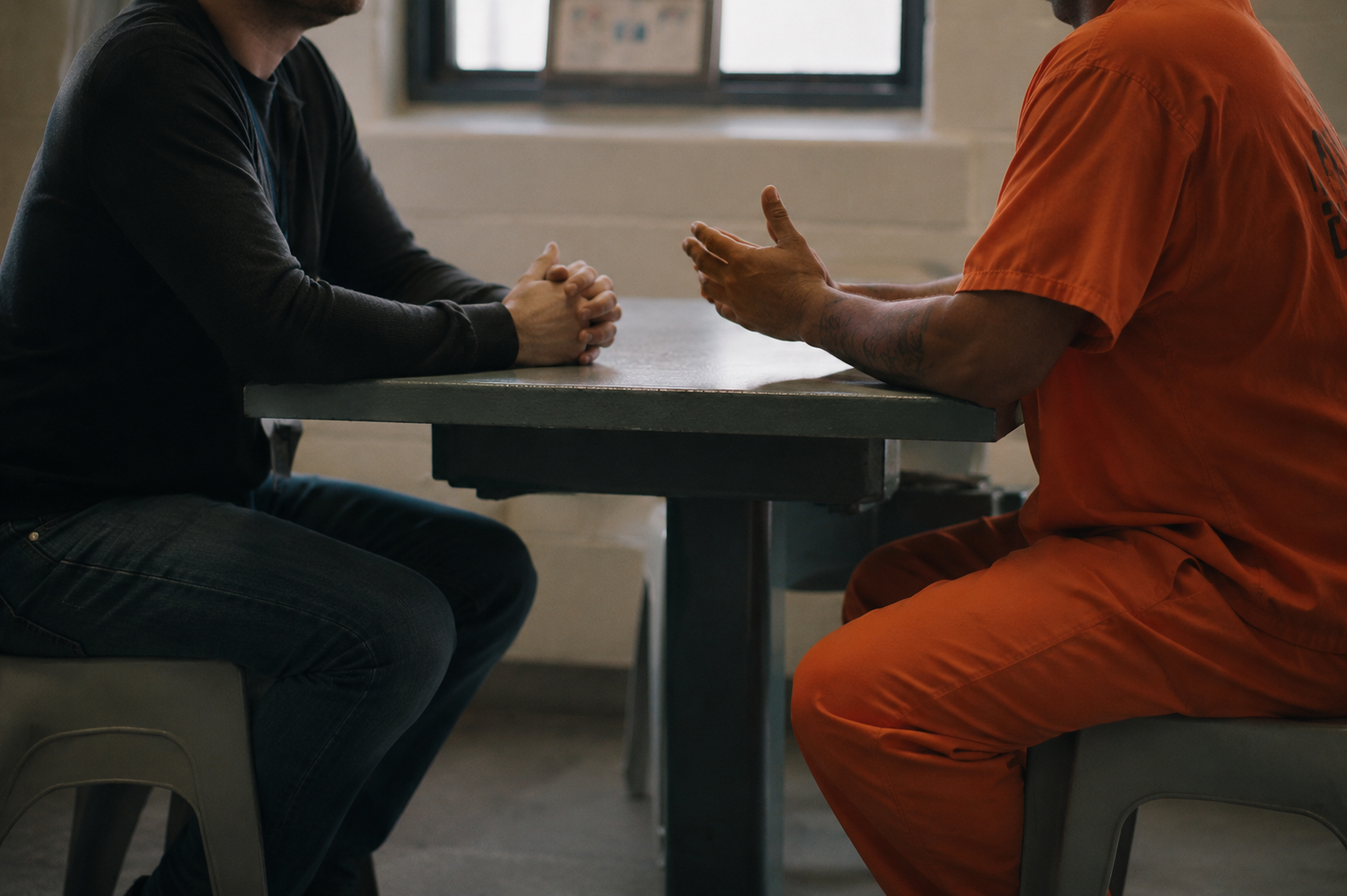 A man in an orange prison uniform talking to a man in casual clothing during an interview in a detention center.