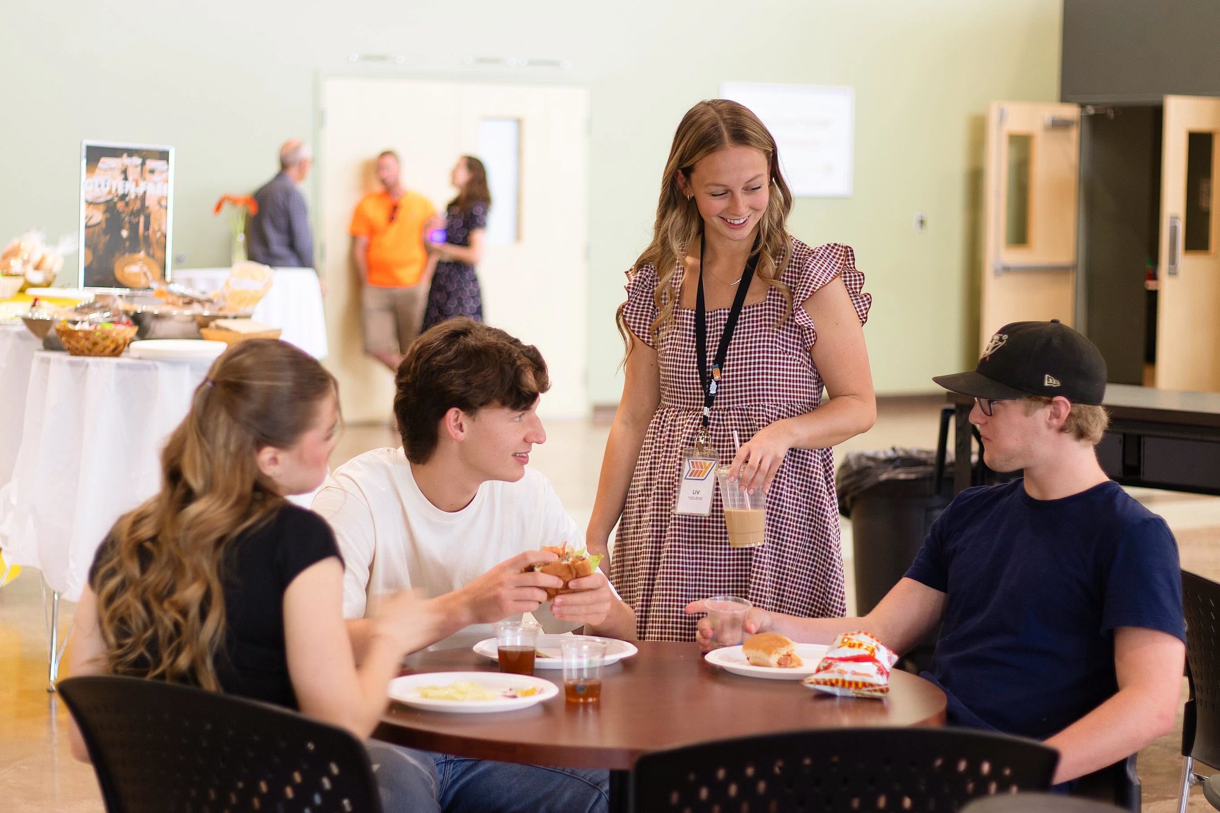 A group of young people sitting around a table in a casual setting, engaging in conversation, with food and drinks on the table. A young woman is standing, smiling, and interacting with the seated individuals.