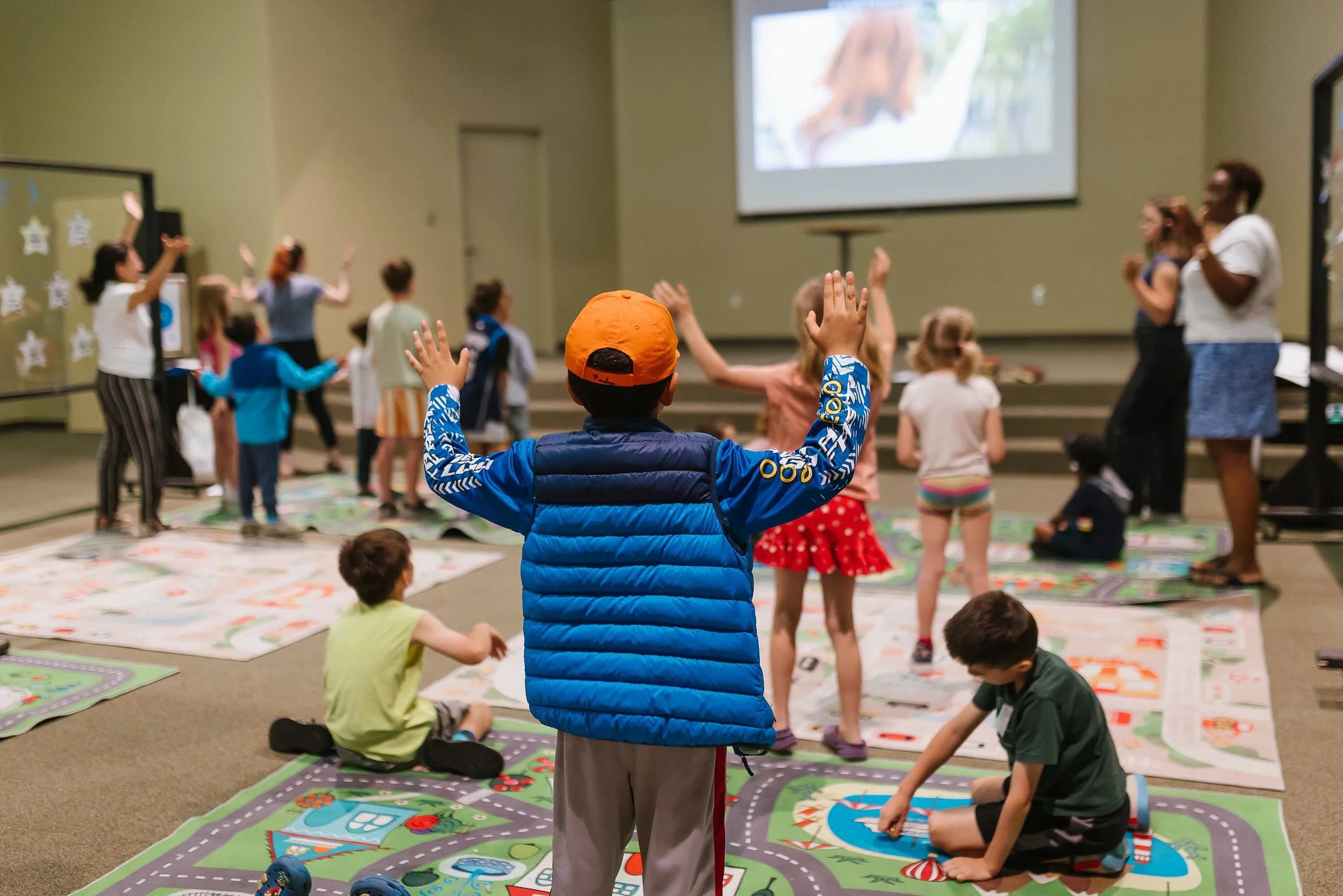 Children and adults participating in an indoor activity with a large game board on the floor, some raising their hands, and a dog projected on a screen