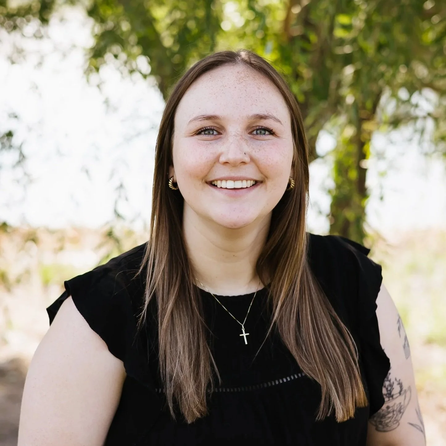 A smiling woman with long brown hair, wearing a black top and a necklace with a cross, standing outdoors with lush green foliage in the background.