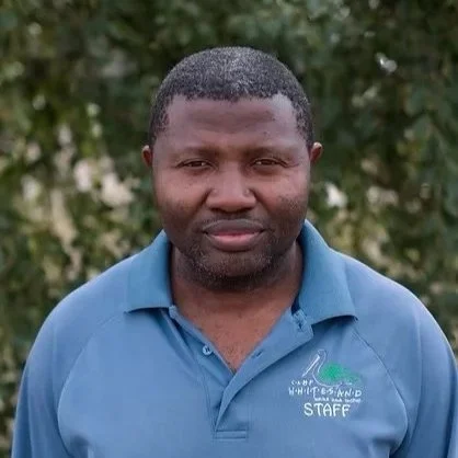 A man with dark hair and skin wearing a blue polo shirt with a logo and the words 'STAFF' on it, standing outdoors with green foliage in the background.