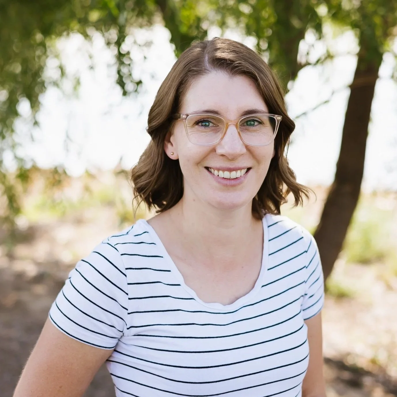 A woman with glasses smiling outdoors with green foliage in the background.