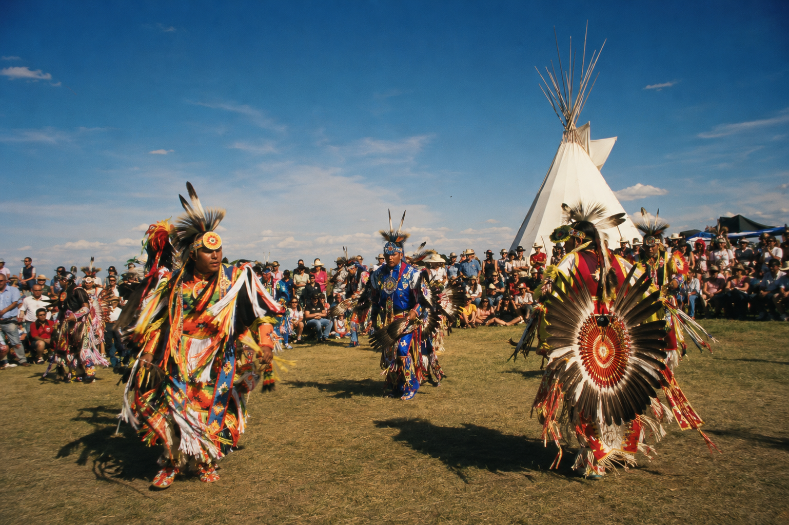 Native American dancers in colorful traditional regalia performing a dance outdoors during a cultural event, with a teepee and crowd watching in the background under a blue sky.