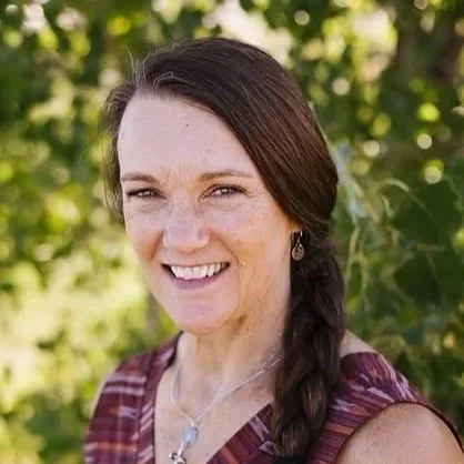 A smiling woman with long dark hair in a braid, wearing a sleeveless striped top and jewelry, standing outdoors with green foliage in the background.