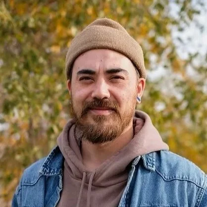 A man with a beard and mustache wearing a tan beanie and gray hoodie underneath a blue denim jacket, standing outdoors with autumn-colored trees in the background.