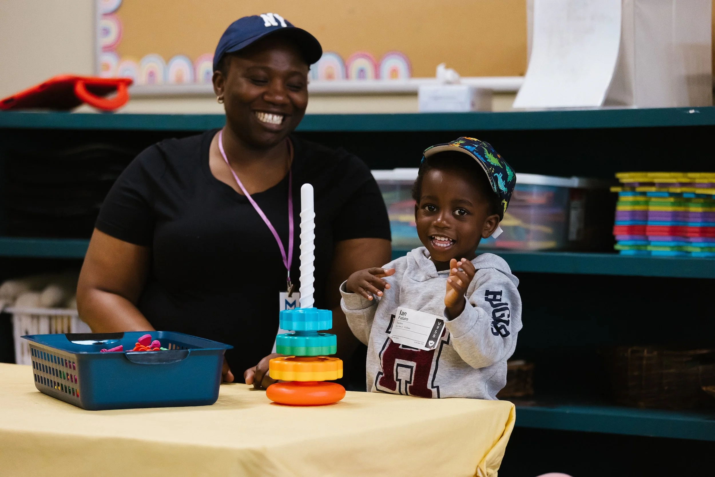 A woman and a young boy enjoying a game of Jenga at a table in a classroom or playroom. The woman is smiling, and the boy is excited as he prepares to make a move. There are colorful stacking blocks and a blue plastic basket filled with small toys on the table.
