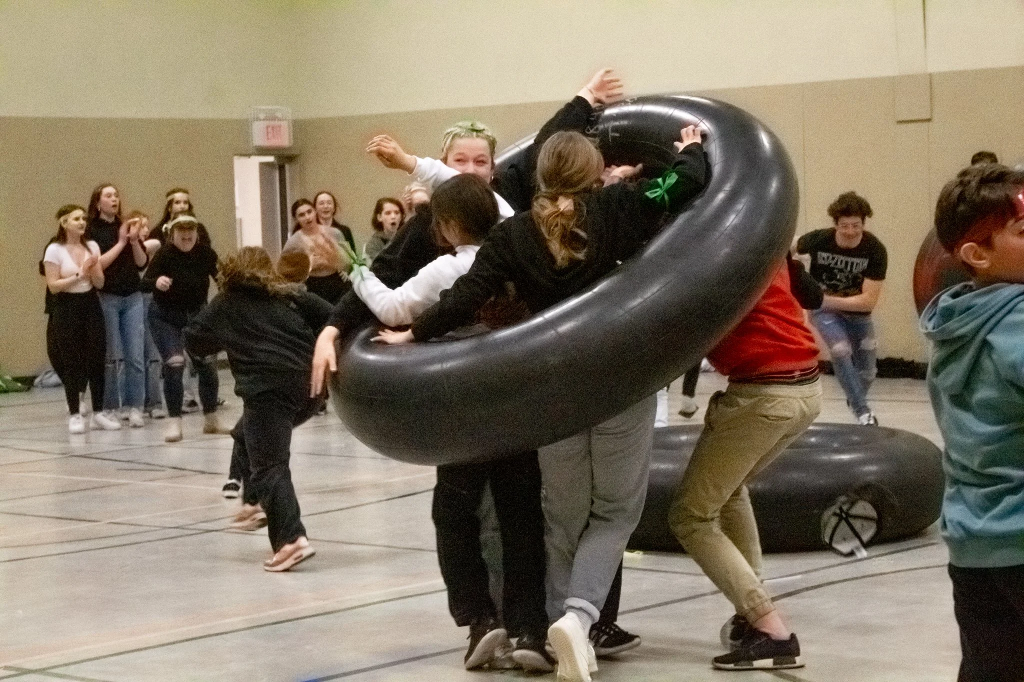 Students in a gymnasium participating in a human foosball game involving a large inflatable ring, with onlookers and clapping peers in the background.