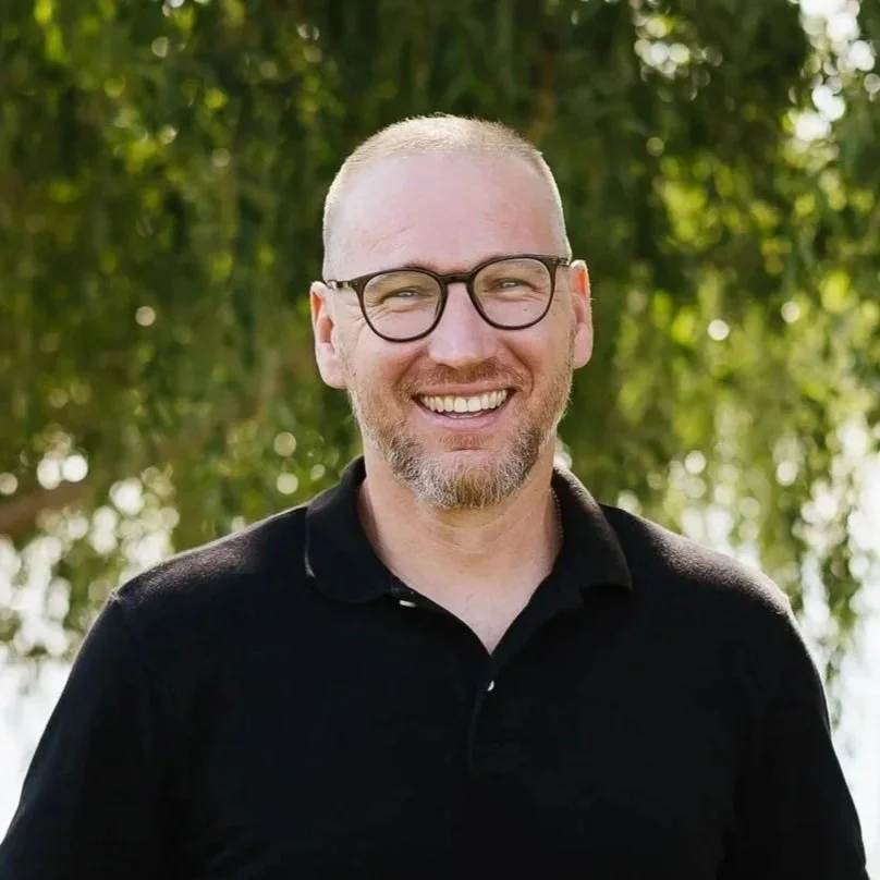 Smiling man with a beard and short hair, wearing a white shirt, outdoors with green foliage in the background.