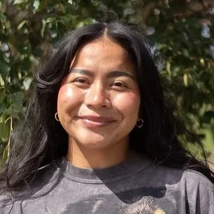 A woman with long black hair smiling outdoors with greenery in the background.