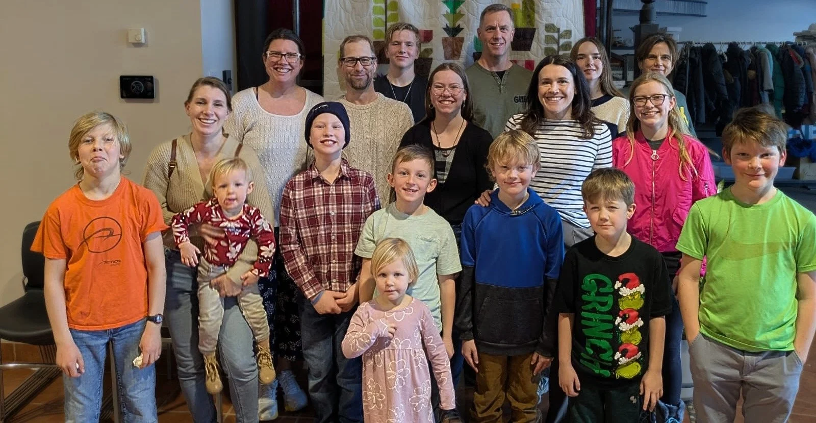 A group of children and adults posing together indoors, smiling for the photo, with a quilt hanging on the wall behind them.