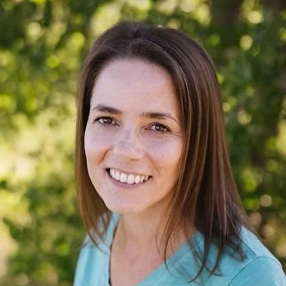 A woman with shoulder-length brown hair, smiling, standing outdoors with green foliage in the background.