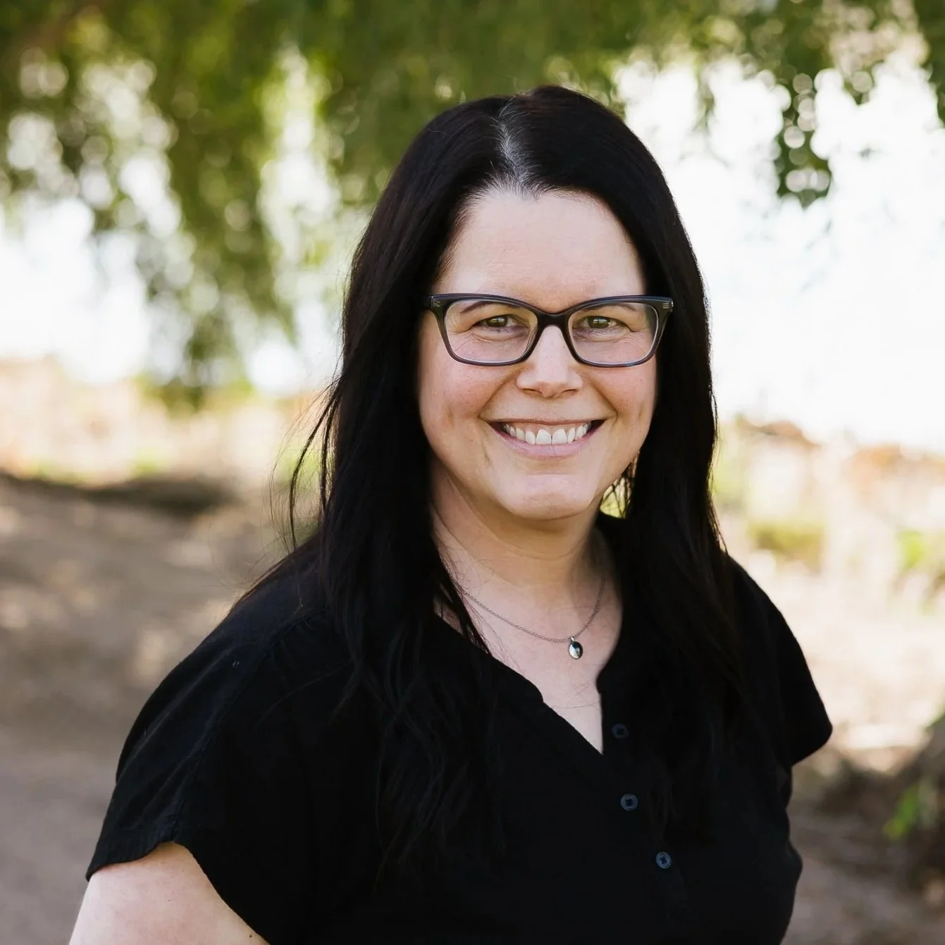 Portrait of a woman with dark wavy hair, glasses, smiling, outdoors with trees in the background.
