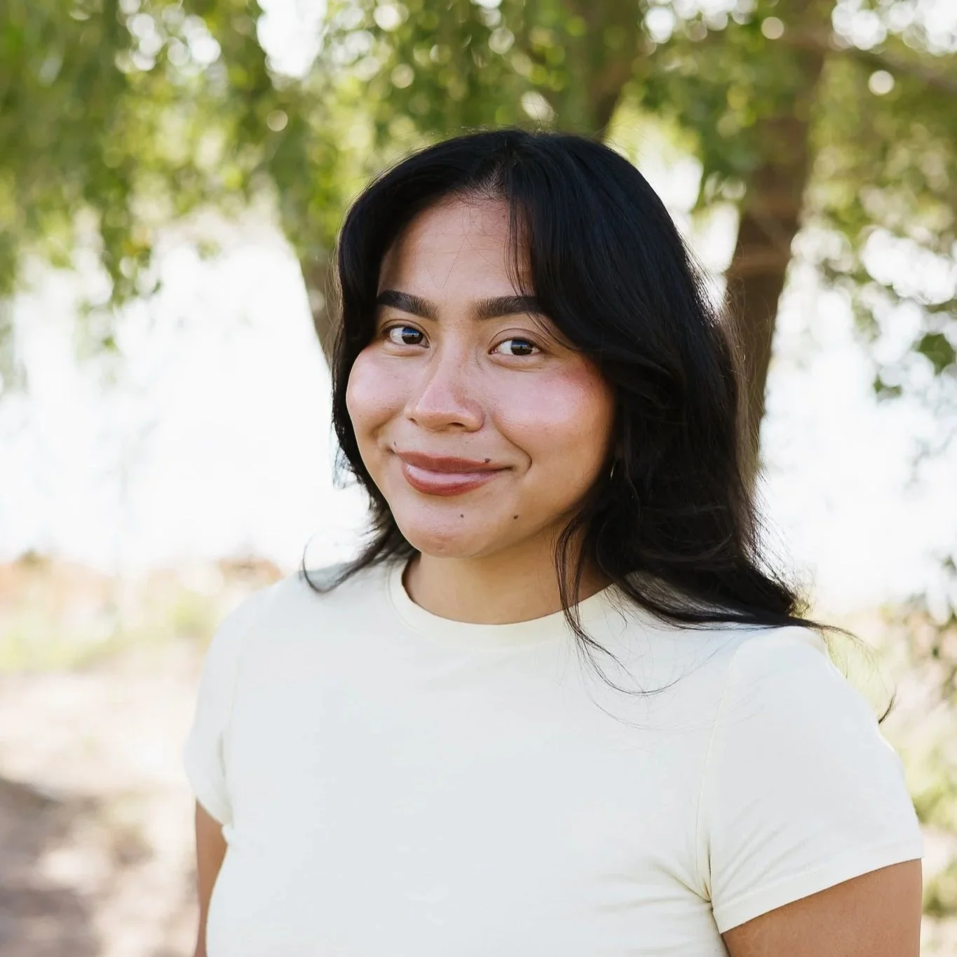 A woman with long black hair smiling outdoors with greenery in the background.