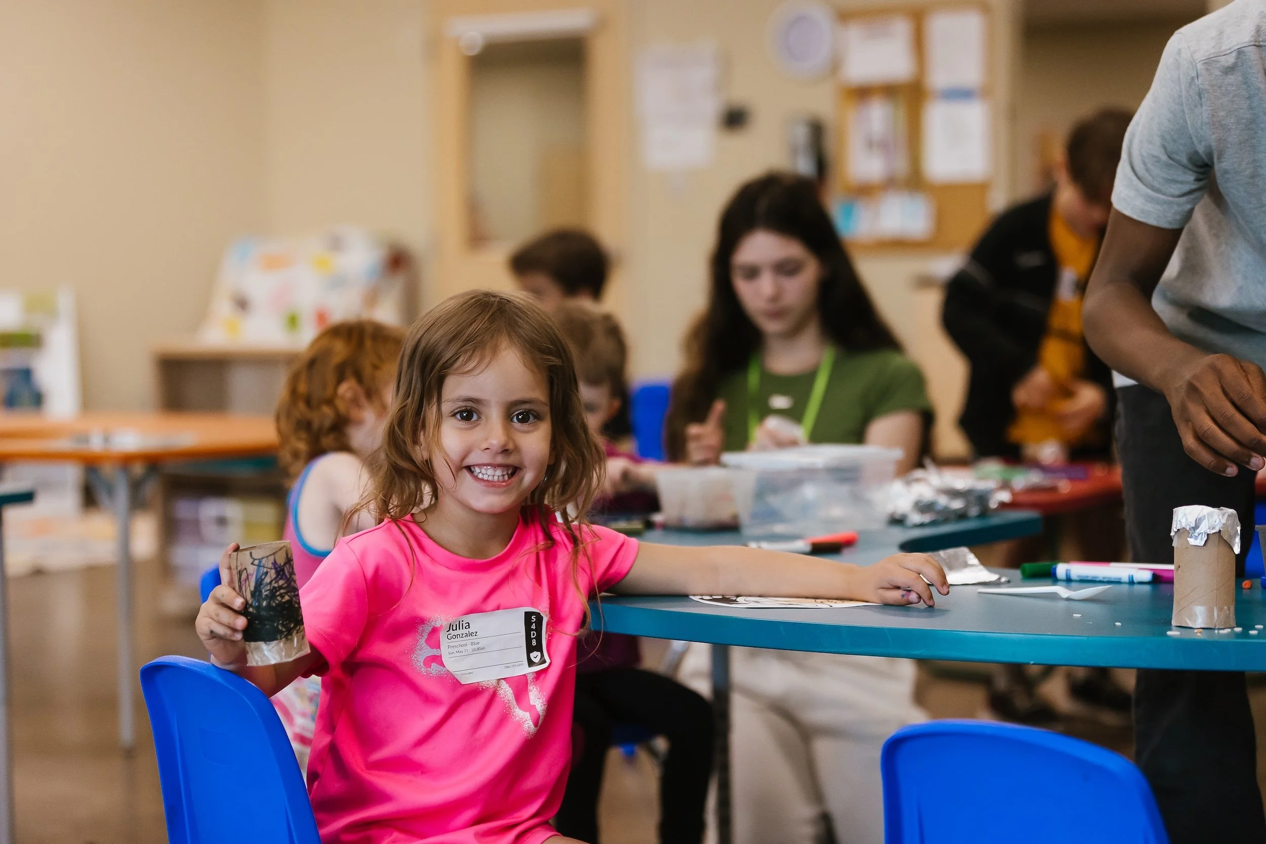 A young girl with brown hair and a bright pink shirt is smiling and holding a decorated cup in a classroom setting. Other children and an adult are in the background, engaging in activities at tables.