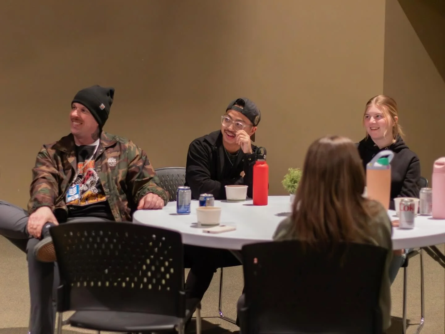 Group of four young adults sitting around a white table, smiling and talking, with a plain beige wall in the background.