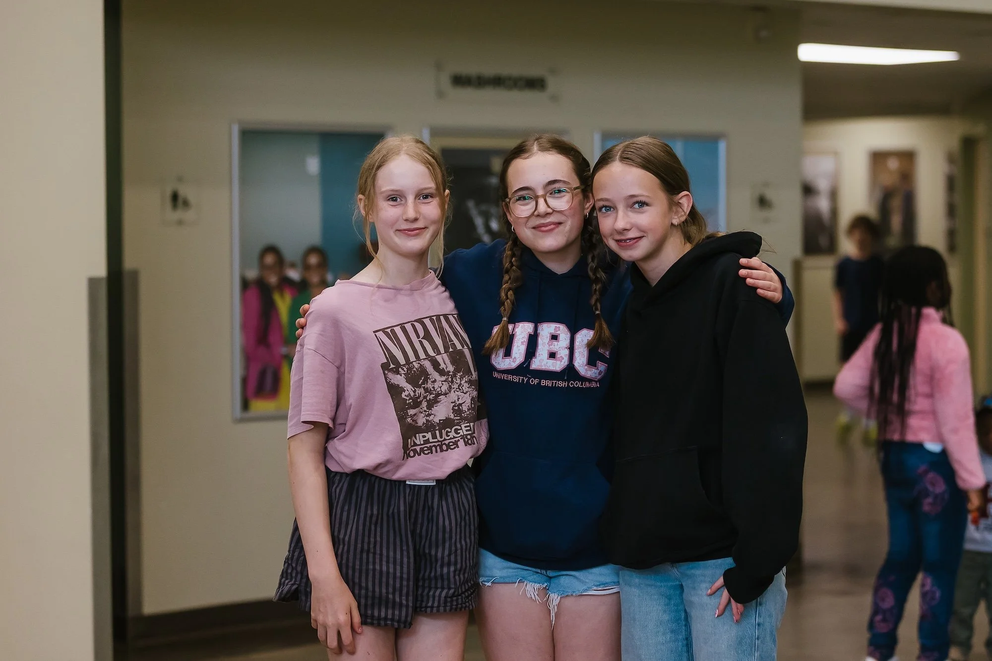 Three young girls standing together in a school hallway, smiling at the camera, with other children blurred in the background.