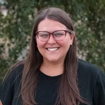 Portrait of a smiling woman with long brown hair, glasses, wearing a black shirt, outdoors with greenery in the background.