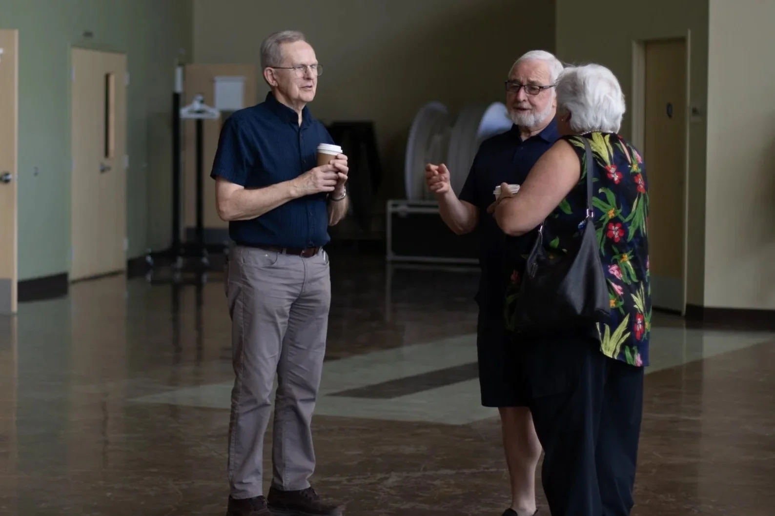 Three elderly people, two men and one woman, standing and talking in a room with wooden floors and light green walls. The men are holding coffee cups, and the woman has a colorful floral top and a handbag.