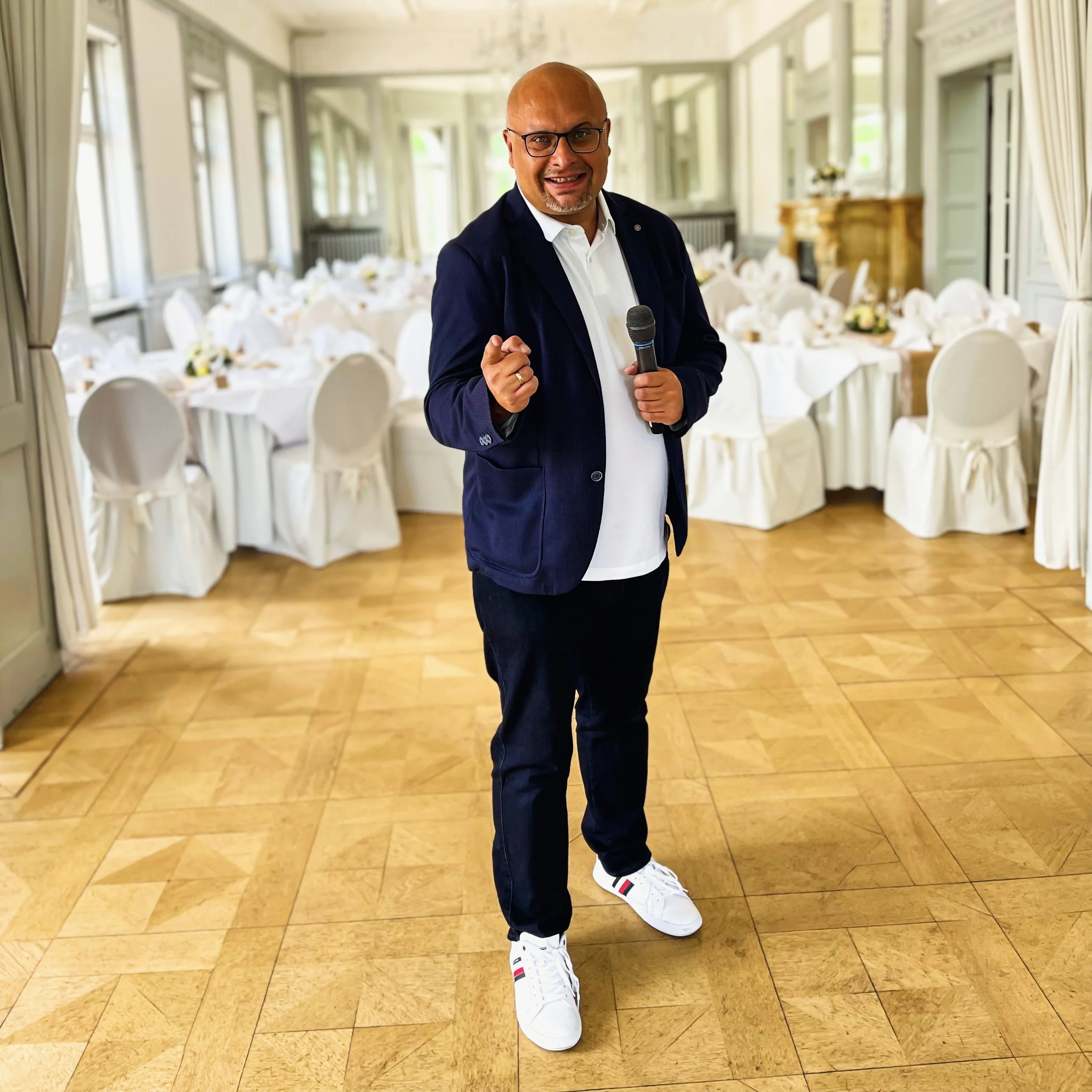 Ein Mann mit Brille, dunklem Jackett, weißem Hemd, Turnschuhen und einem Mikrofon in der Hand steht vor einer festlich gedeckten Tafel in einem großen, elegant eingerichteten Raum.