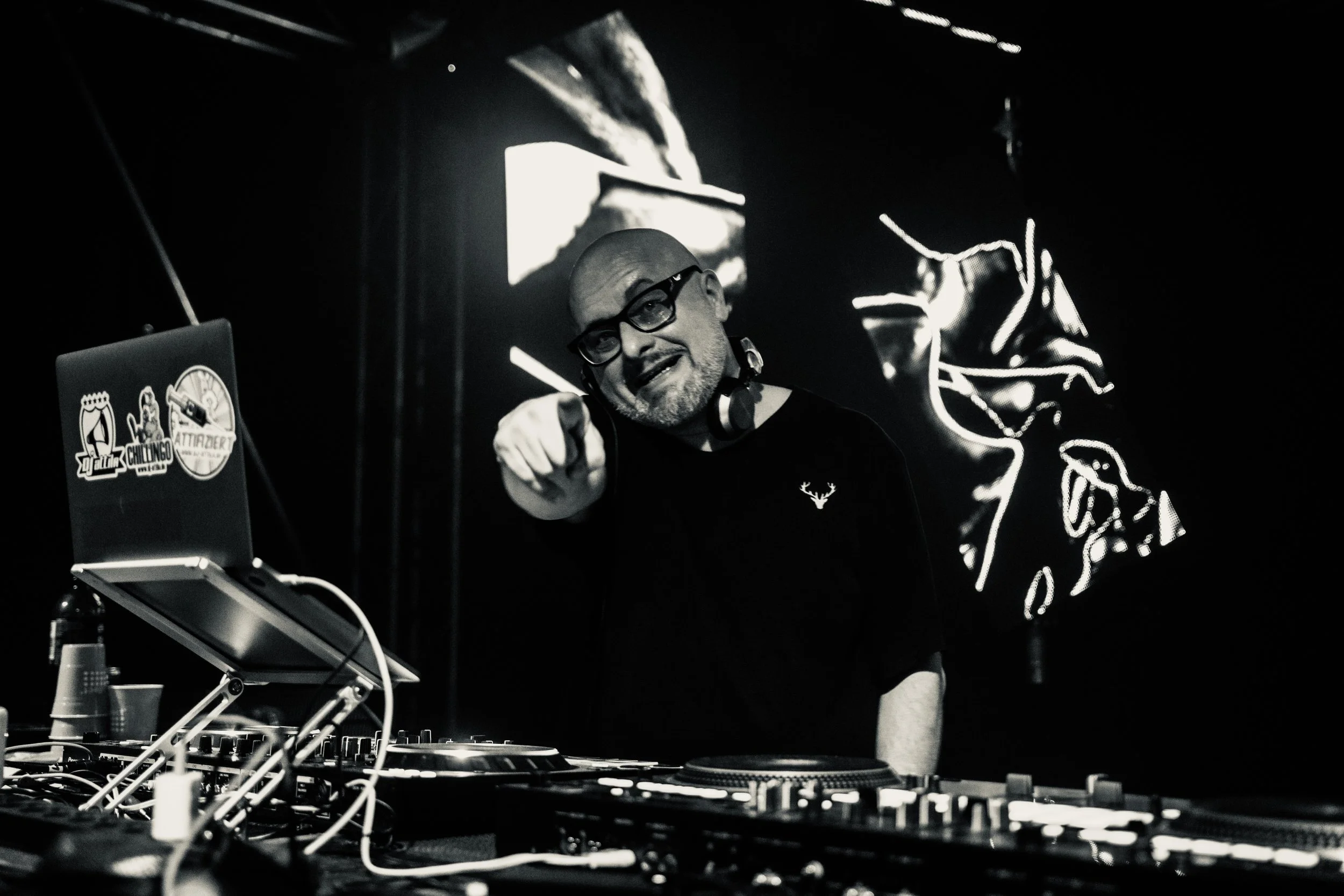 Ein DJ mit Kopfhörern hinter einem DJ-Mischpult, umgeben von elektronischer Ausrüstung, in einem dunklen Club oder Veranstaltungsraum.