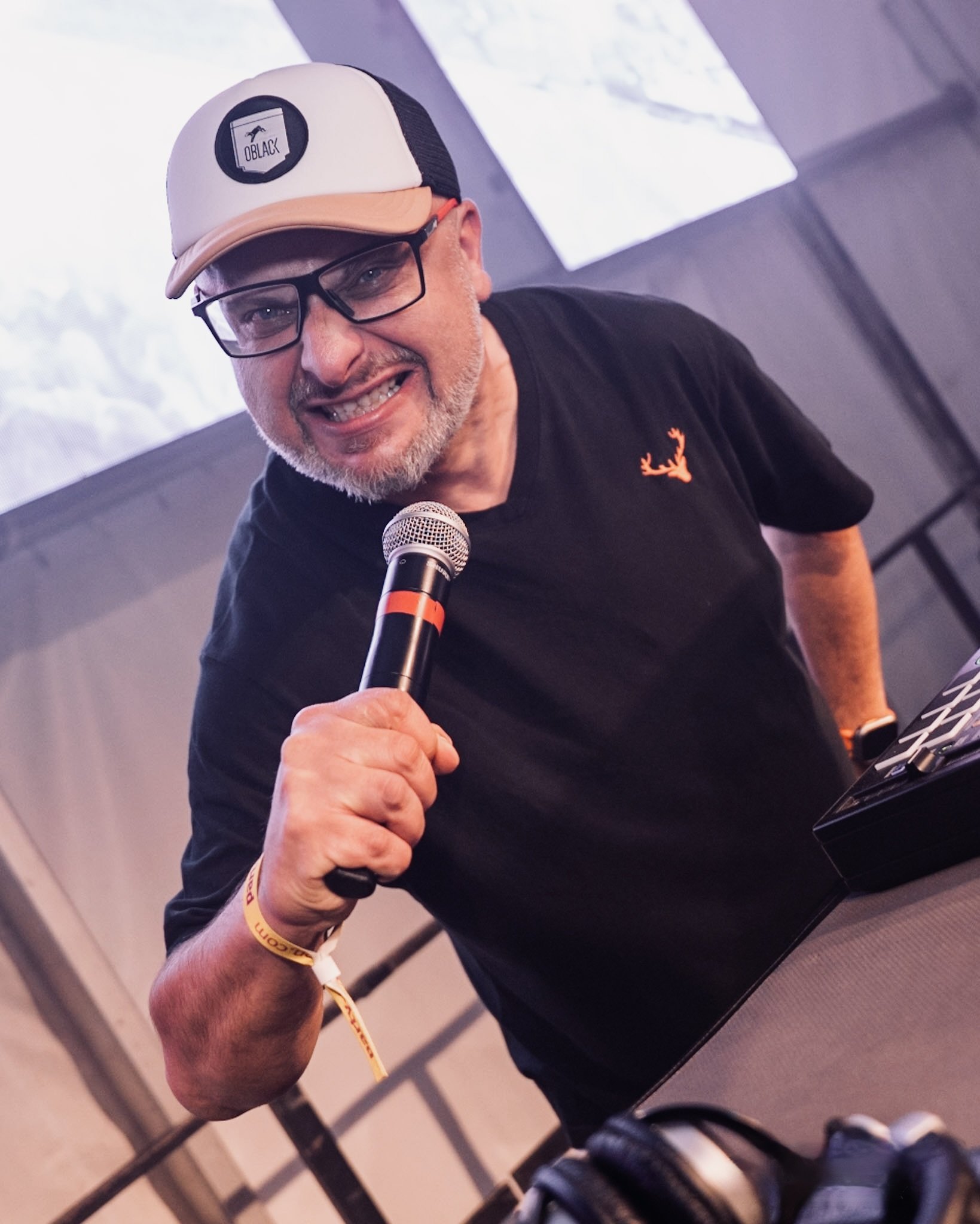 DJ Attila aus Heidenau trägt eine blaue T-Shirt mit Hirsch-Logo, eine weiße Cap mit schwarzen Seiten und ein Mikrofon. Er lächelt und zeigt mit seiner Hand auf die Kamera. Er ist auch buchbar als DJ in Dresden, Pirna, Glashütte, Freital