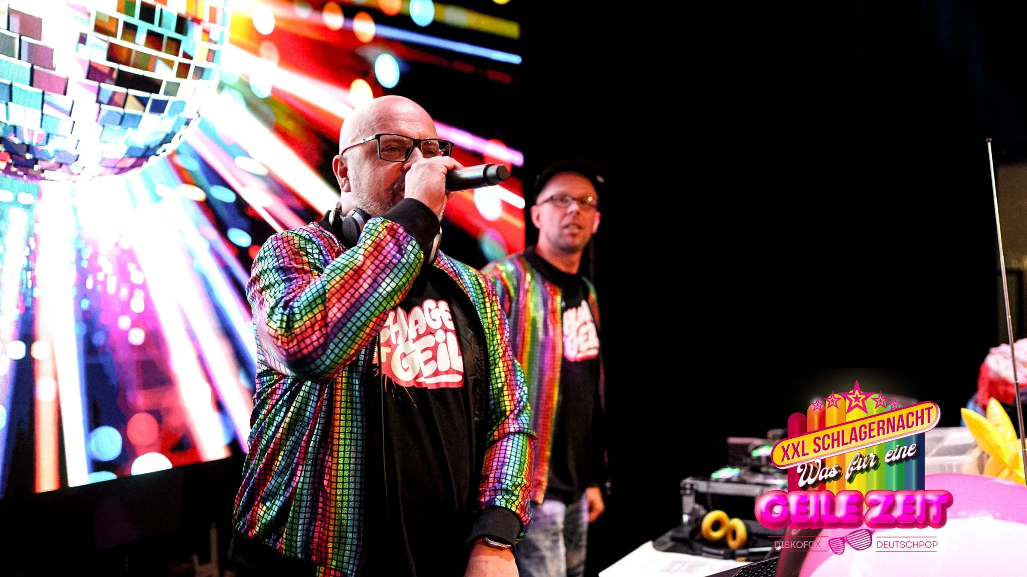 Zwei Männer bei einer DJ-Performance auf einer bunten Party, beide tragen knallbunte, regenbogenartige Jacken, im Hintergrund bunte Lichter und eine Discokugel.