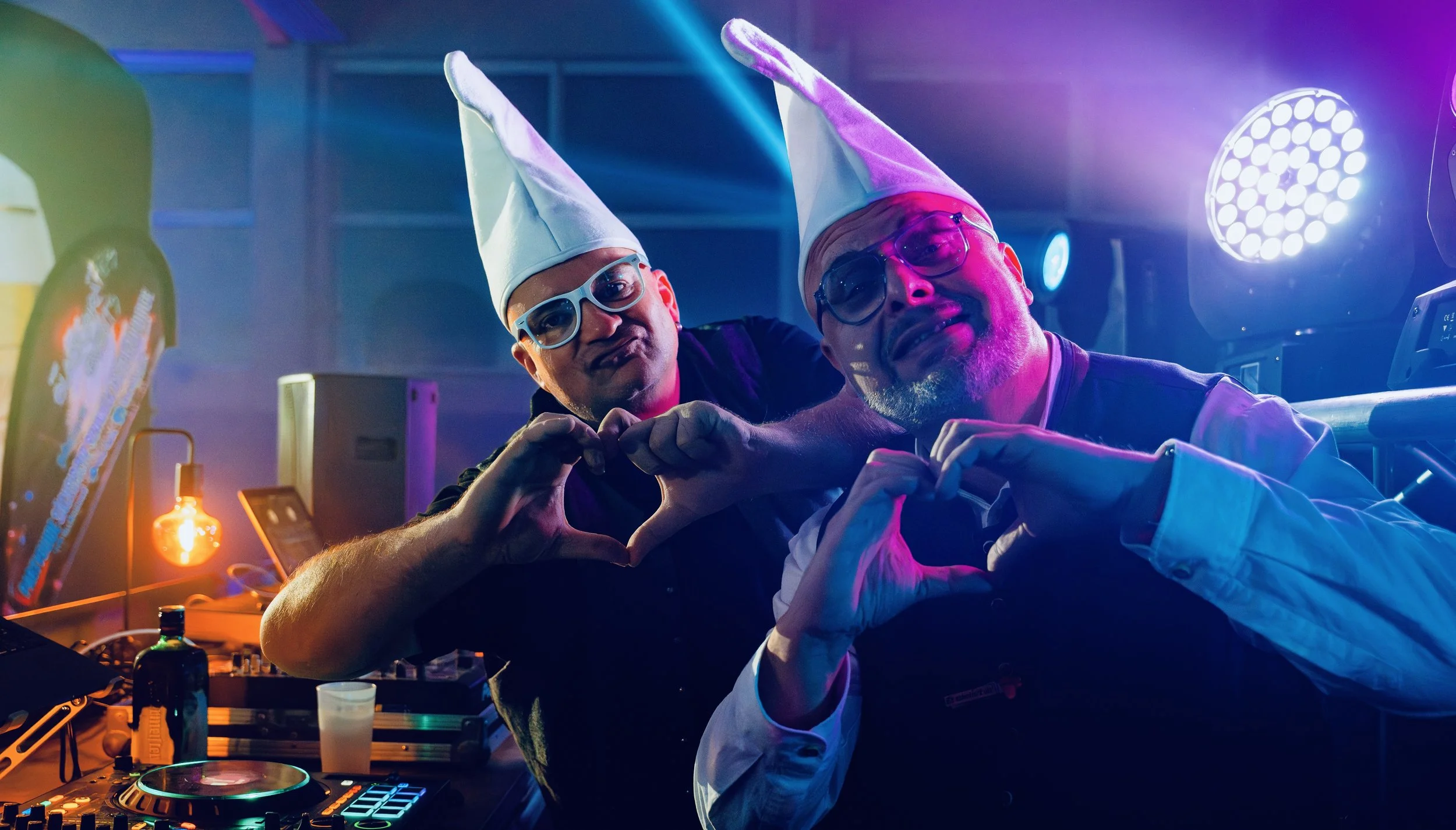 Zwei Männer bei einer Party tragen lustige Hüte und Sonnenbrillen, machen ein Herzzeichen mit den Händen und posieren vor DJ-Equipment bei bunter Lichtbeleuchtung.
