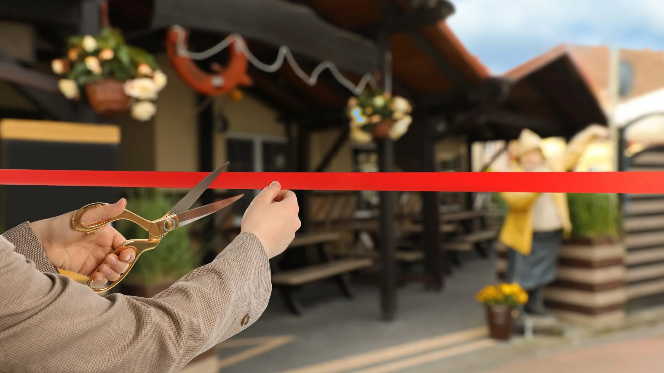 Person in a beige blazer cutting a red ribbon with gold scissors at an outdoor event.
