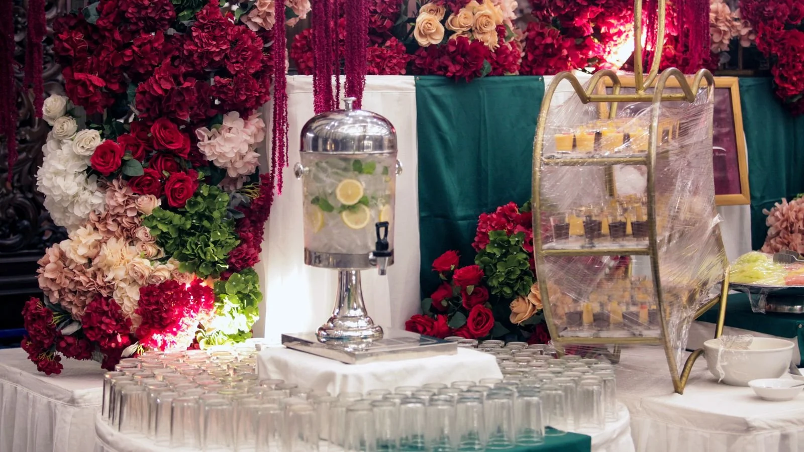 A beverage dispenser with lemon slices and mint leaves, a ladder with small cups covered in plastic wrap, and a floral backdrop of red, pink, white, and peach roses at a decorated event table.