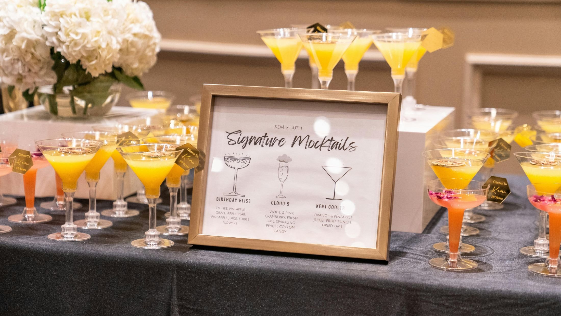 A display of yellow and pink cocktails in martini glasses with a framed menu sign in the foreground and white floral arrangement in the background.