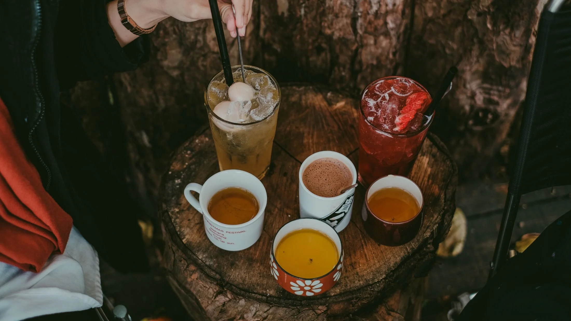A top-down view of a rustic wooden table with multiple beverages, including two tall glasses with ice and straws, and several cups of tea or coffee. One of the glasses contains a yellow drink with ice, and the other has a red drink with ice. The cups and glasses are placed against a dark, textured background.