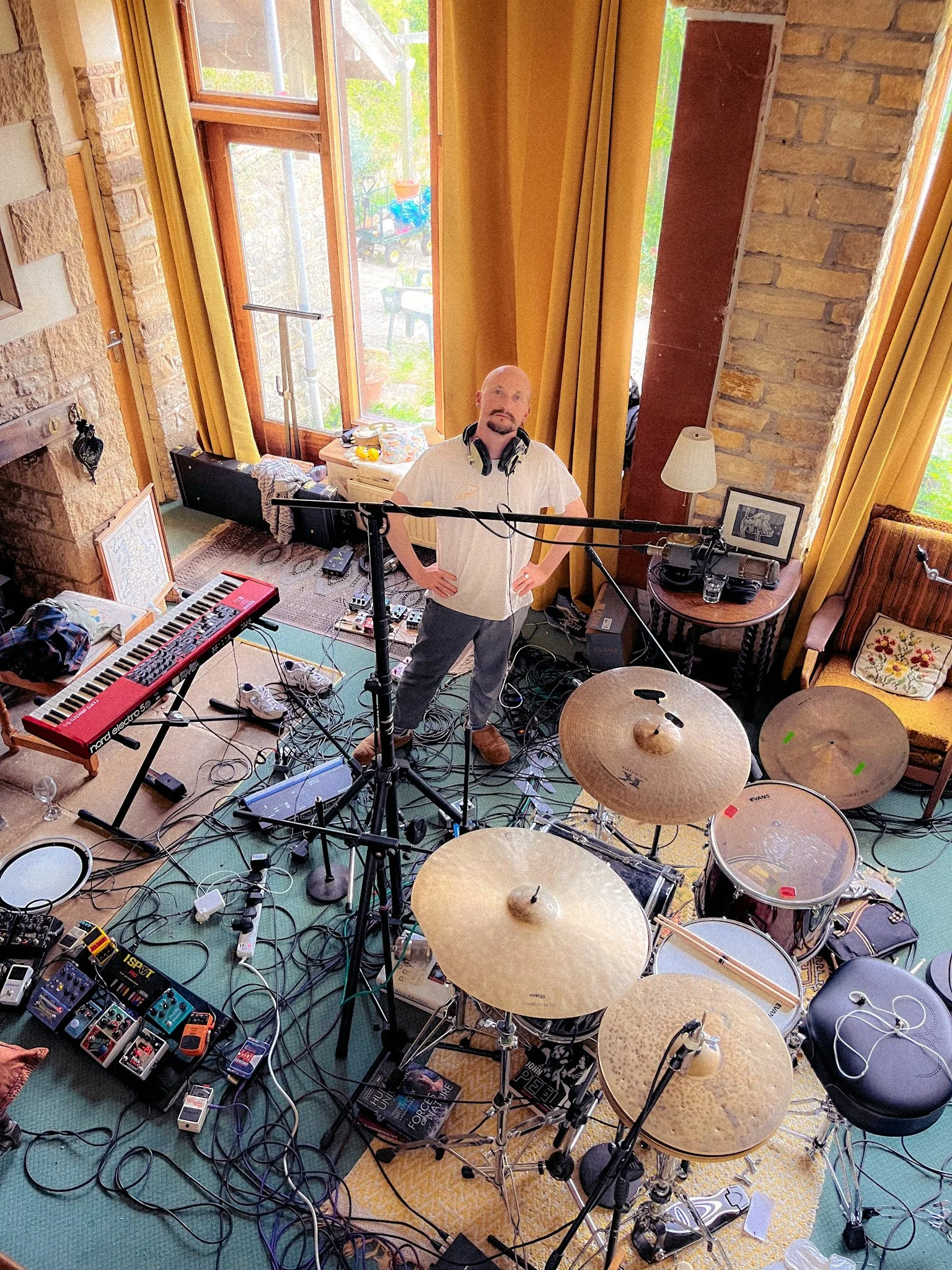 A man standing in a music studio with musical instruments, including a drum set, keyboard, and guitar pedals, surrounded by cables, books, and equipment, with large windows and yellow curtains behind him.
