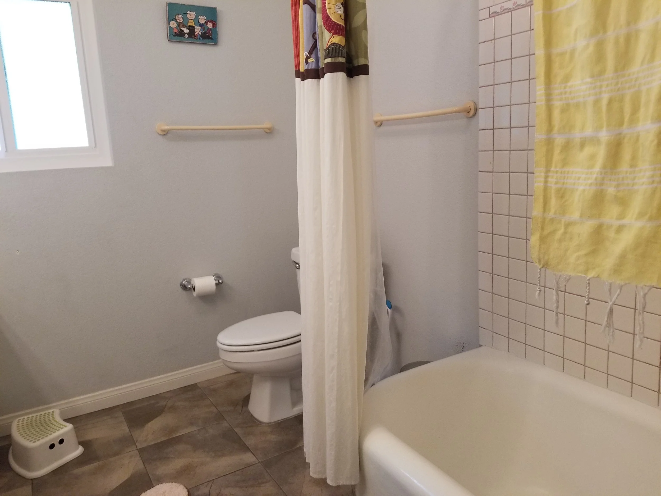 A small bathroom with a toilet, shower with a yellow and white striped towel, window, and wall art featuring cartoon characters.