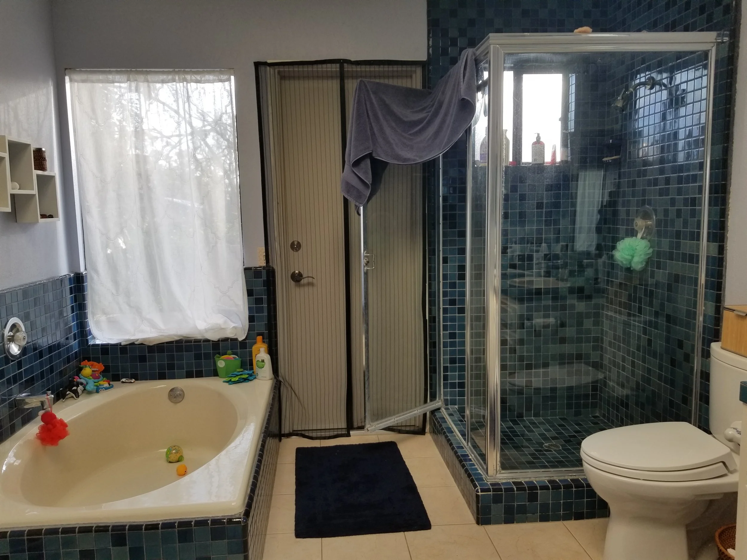 A bathroom with a bathtub, a shower enclosure, a toilet, and a window with a curtain. The bathtub has toys and a red bath sponge, and the shower area contains bottles of shampoo and body wash.