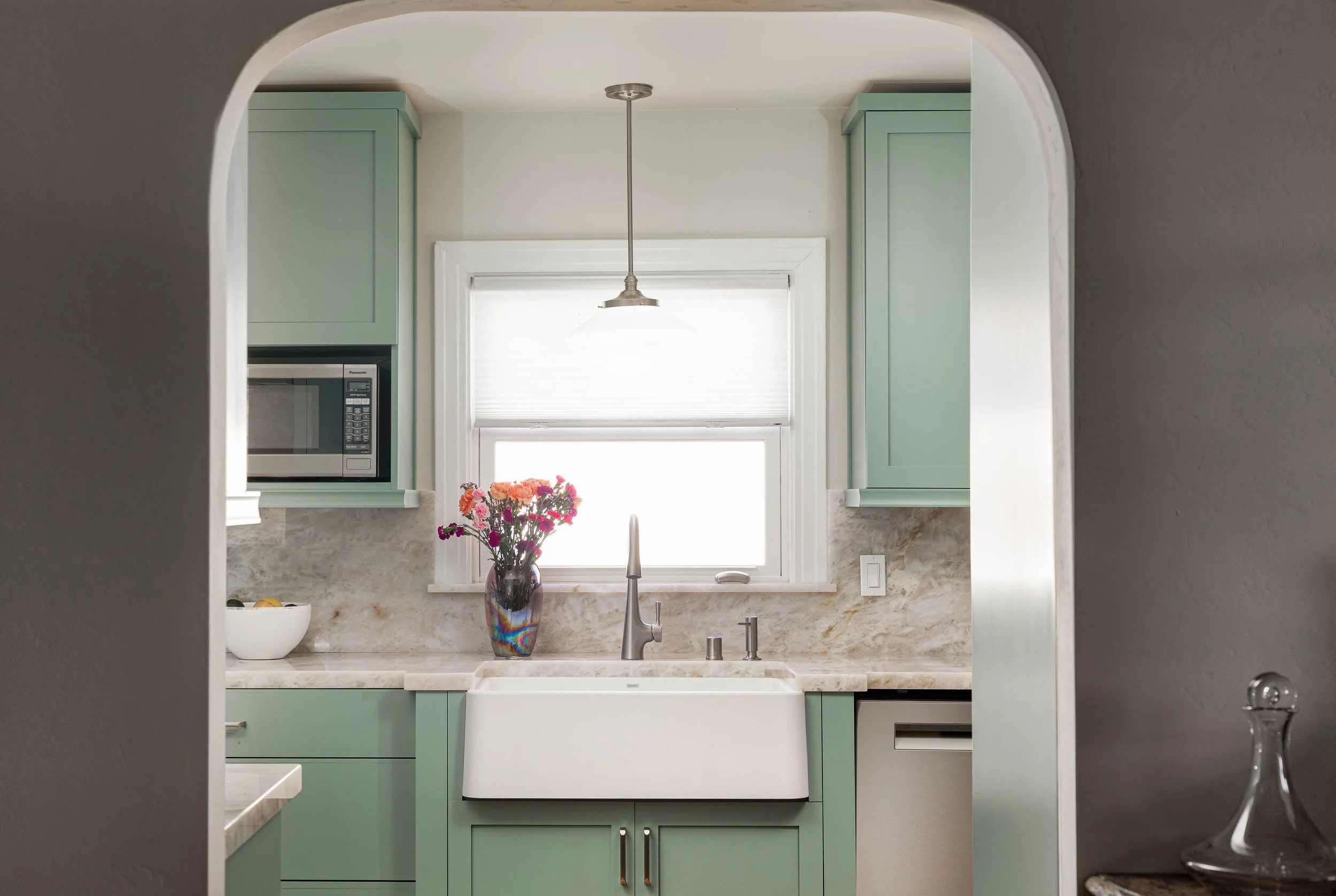 View of a kitchen seen through an arched doorway with green cabinets, a marble countertop, a window with white blinds, a vase with flowers, and a microwave oven.
