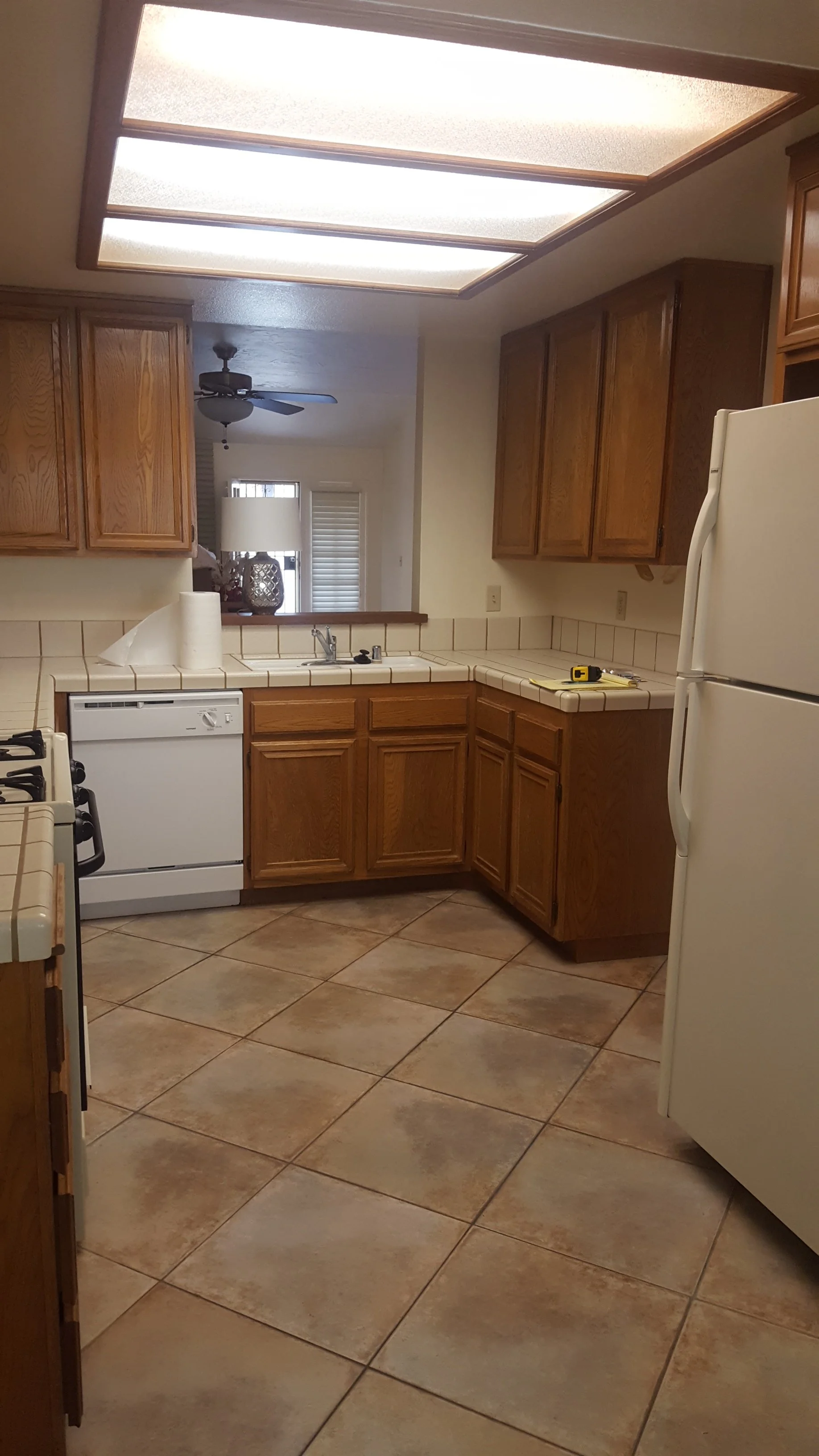 Kitchen with wooden cabinets, white appliances, tiled counters and floor, ceiling light, and a view into a living room with a ceiling fan and window.