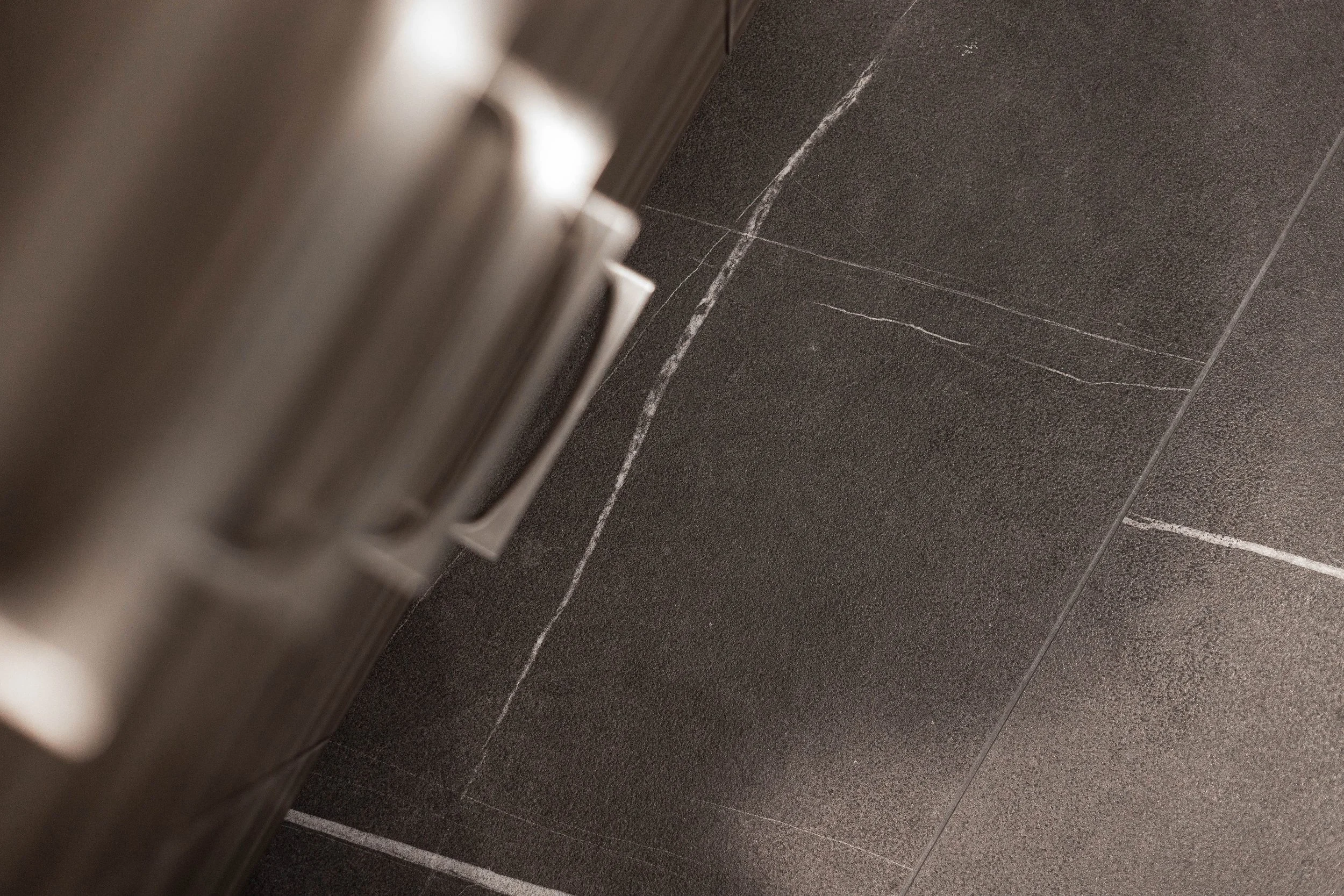 Close-up of a section of black tiled floor and a silver-colored metal surface, possibly part of a bathroom or elevator interior.