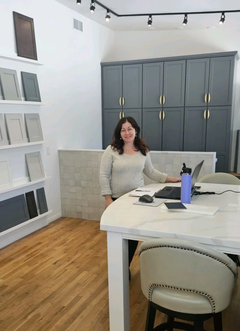 A woman with dark hair smiling at a desk in a room with gray cabinets and sample paint color swatches on the wall.