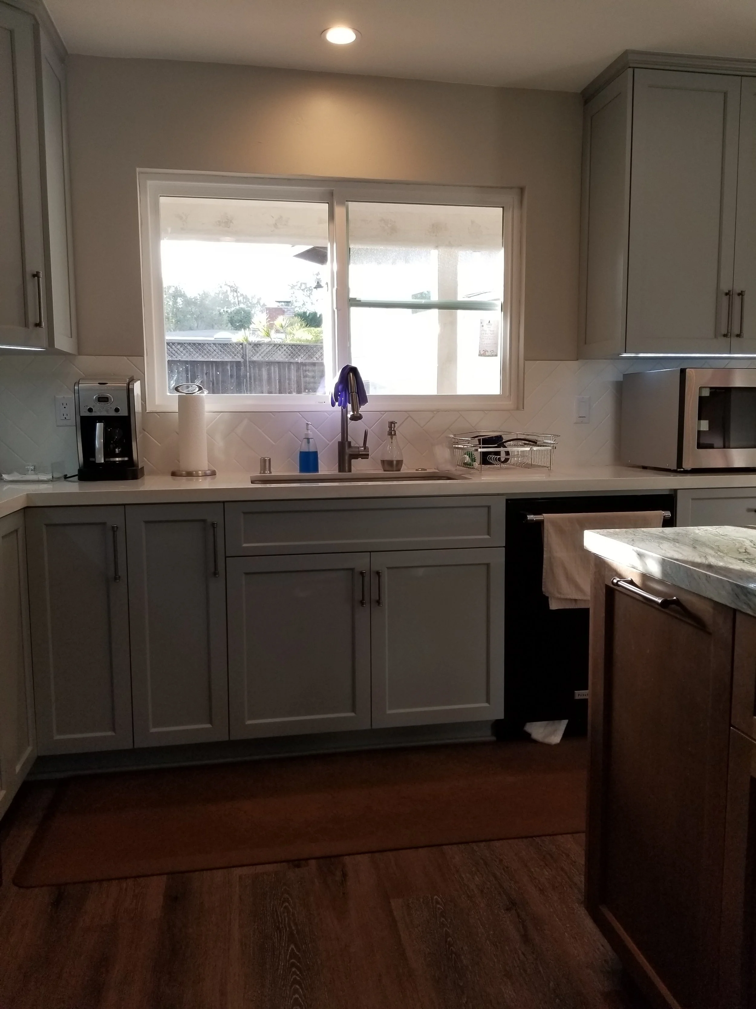 A kitchen with gray cabinets, a window above the sink, a coffee maker, paper towels, a soap dispenser, a drying rack, a microwave, and a towel on an oven handle, with wooden flooring and a rug in front of the sink.